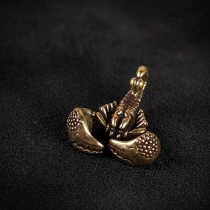 May include: A detailed bronze-colored scorpion figurine with prominent claws and a segmented body. The scorpion is posed with its tail curved upwards, showcasing its stinger. The figurine is set against a dark background.