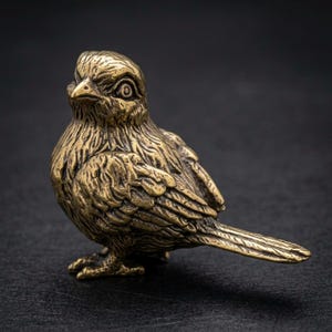 Puede incluir: Una detallada figura de pájaro de bronce con plumas texturizadas y una mirada enfocada. La pequeña escultura está en posición vertical, mostrando sus delicadas patas y cola. El pájaro es un objeto decorativo.