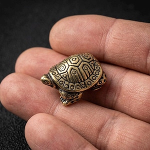 Könnte beinhalten: Kleine, detaillierte Schildkrötenfigur aus Messing. Die Schildkröte hat einen strukturierten Panzer mit geometrischen Mustern und sichtbaren Beinen und Kopf. Die Figur wird in einer Hand gehalten, um ihre Größe und ihr kompliziertes Design zu zeigen. Dekorativer Artikel.