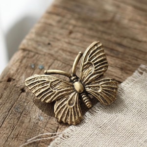 May include: A detailed, antique-style butterfly brooch in a warm, brassy gold tone. The wings and body are intricately designed with textured details. The brooch is resting on a weathered wooden surface with a linen fabric in the background.