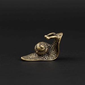 May include: A small, detailed brass snail figurine. The snail has a textured body and a spiraled shell. The figurine is a warm, antique gold color and sits on a dark background.