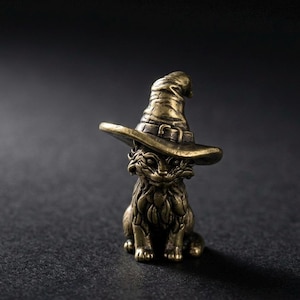 Könnte beinhalten: Bronze-Katzenfigur mit Hexenhut. Die Katze sitzt aufrecht mit den Pfoten nach vorne. Der Hut hat eine breite Krempe und eine Schnalle. Die Figur hat eine detaillierte, strukturierte Oberfläche.