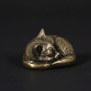 Peut inclure: Petite sculpture en bronze représentant un chat enroulé en position de sommeil. Le pelage du chat est détaillé avec des lignes gravées, et ses oreilles et sa queue sont visibles. La sculpture est posée sur un fond sombre.