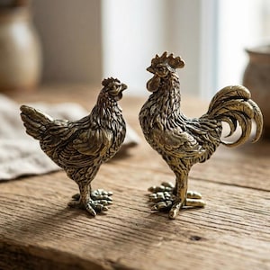 Könnte beinhalten: Zwei bronzefarbene Hühnerfiguren. Eine ist eine Henne, die andere ein Hahn, beide mit detaillierten Federstrukturen und stehen auf einer Holzoberfläche. Der Hahn hat einen großen, gebogenen Schwanz und einen ausgeprägten Kamm.