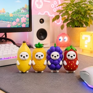 May include: Four small animal figurines standing in a row, each wearing a different fruit-shaped costume. The costumes are a yellow banana, a yellow pineapple, a cluster of purple grapes, and a red strawberry with green leaves on top.