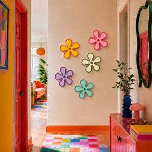Puede incluir: Cinco decoraciones de pared con forma de flor en colores naranja, rosa, verde claro, morado y azul cerceta están dispuestas sobre una pared beige. Cada flor presenta un recorte circular central y pétalos suaves y redondeados.