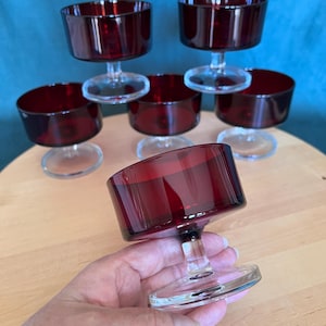 May include: A set of six dessert cups featuring deep red glass bowls and clear glass stems and circular bases. The cups have a wide, cylindrical bowl shape with a smooth rim. One cup is held in the foreground, while five others are arranged behind.