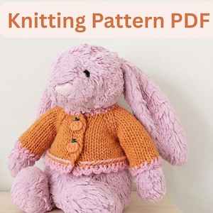 Pode incluir: Um coelho de pelúcia lavanda usando um cardigã laranja de malha com botões em forma de abóbora e acabamento rosa. A imagem inclui o texto "Knitting Pattern PDF". O coelho é um brinquedo de pelúcia.