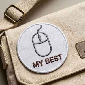 May include: A round, white embroidered patch with a gray computer mouse design and the words "MY BEST" in brown lettering. The patch is affixed to a beige canvas bag with a shoulder strap and metal buckle.