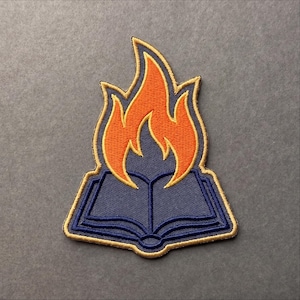 May include: An embroidered patch featuring an open book in dark blue with a gold border. Rising from the center of the book is a stylized flame design in bright orange with a gold outline, all set against a dark blue background.