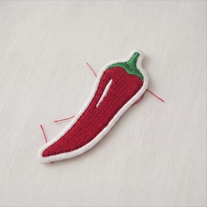 May include: A chili pepper patch featuring red embroidery for the body, a green stem, and a white border. The design includes a small white highlight line on the pepper. Several loose red threads extend from the edges of the embroidered patch.