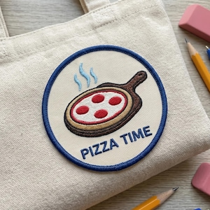 May include: A beige tote bag with a circular patch featuring a pizza on a wooden paddle. The pizza has red pepperoni and steam rising from it. The patch is outlined in blue, with the words "PIZZA TIME" in blue.