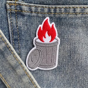 May include: An embroidered patch featuring a gray trash can with a detached lid and bright red flames rising from the top. The design is outlined in white thread and is attached to a blue denim fabric surface with visible stitching and seams.