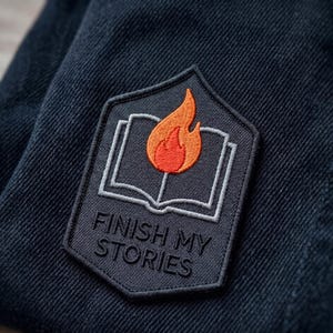May include: A dark gray embroidered patch with the text "FINISH MY STORIES". The patch features a burning flame above an open book. The patch is sewn onto a dark blue denim fabric.