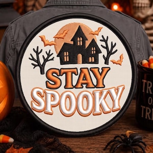 May include: A circular embroidered patch featuring a black haunted house silhouette against an orange moon, surrounded by bare trees and bats. The text STAY SPOOKY is stitched in orange and white letters on a cream background with a black border.