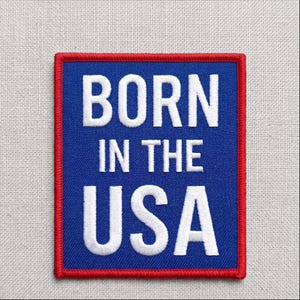 May include: Embroidered patch with the text "BORN IN THE USA" in white capital letters on a blue background. The patch has a red border and is square-shaped.