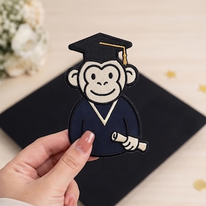 May include: An embroidered patch featuring a cartoon monkey wearing a black graduation cap with a gold tassel and a navy blue graduation gown. The monkey is holding a rolled-up diploma in one hand, rendered in cream and black thread colors.