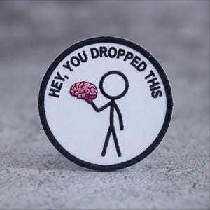 May include: A circular embroidered patch with a white background and a black border. It features a black stick figure holding a pink brain, with the black text "HEY, YOU DROPPED THIS" arched along the top edge of the circular design.