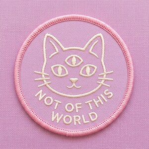 May include: A circular embroidered patch featuring a light pink background with a cream-colored outline of a cat head with three eyes. The text "NOT OF THIS WORLD" is embroidered in cream below the cat head, and the patch has a stitched pink border.
