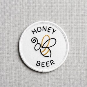 May include: White, circular embroidered patch with the words "HONEY" and "BEER" in black. A stylized bee with black and gold details is in the center. The patch is likely for clothing or accessories.