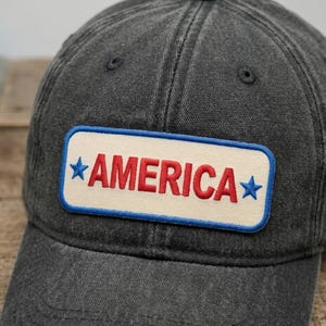 May include: A dark gray baseball cap with a rectangular patch. The patch has a white background with the word "AMERICA" in red letters, framed by a blue border and blue stars.