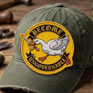 May include: An olive green baseball cap featuring a circular embroidered patch. The patch has a yellow background with a black border and text that reads BECOME UNGOVERNABLE. A white goose holding a silver sword in its beak is centered.