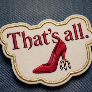 May include: An embroidered patch with a cream-colored base and gold-stitched scalloped edges. It features the text "That's all." in dark red thread, accompanied by an illustration of a red high-heeled shoe with a gold trident-shaped heel.
