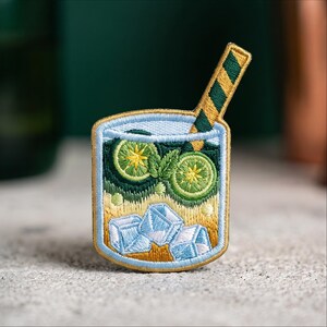 May include: An embroidered patch shaped like a glass of cocktail featuring two green lime slices, mint leaves, and light blue ice cubes. The drink is yellow and light blue with a striped yellow and dark green straw extending from the top right.