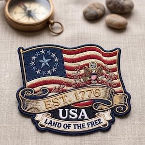May include: An embroidered patch featuring a stylized American flag with a blue canton containing a large star surrounded by smaller stars. It includes a golden eagle emblem, a banner reading EST. 1776, and text reading USA LAND OF THE FREE.