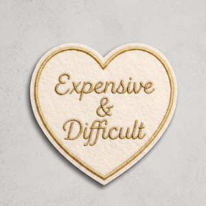 May include: A heart-shaped patch with an off-white felt base and a gold embroidered border. The text "Expensive & Difficult" is embroidered in gold thread in a cursive script across the center of the heart-shaped fabric patch.