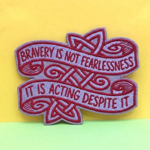 May include: Embroidered patch with the quote "Bravery is not Fearlessness, It is Acting Despite It." The patch features a light purple background with red lettering and decorative Celtic knot designs. The patch is rectangular with a banner shape.