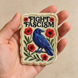 May include: A rectangular embroidered patch with a scalloped edge featuring a blue raven perched on a branch surrounded by red poppy flowers and green leaves. The text FIGHT FASCISM is embroidered in black letters at the top on a cream background.