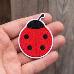 May include: A circular embroidered patch featuring a ladybug design. The patch has a bright red body with four black circular spots, a black head section, and a single black antenna extending from the top, all outlined in a white border.