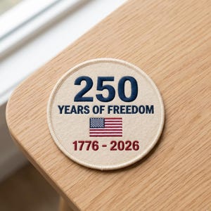 May include: A round patch with the text "250 YEARS OF FREEDOM" and the years "1776-2026". The patch features an American flag design in the center. The number "250" is in navy blue, and the text is in a serif font.
