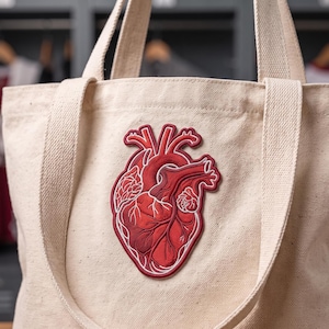 May include: A beige canvas tote bag featuring a large, detailed embroidered patch of an anatomical human heart in shades of red and white. The heart design is centered on the front panel, showcasing intricate stitching of veins and arteries.