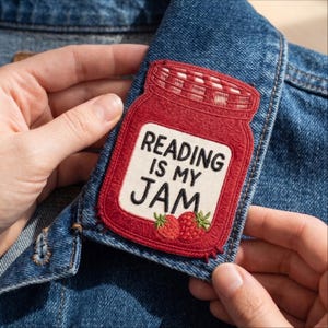 May include: A red embroidered patch shaped like a mason jar attached to a blue denim jacket. The patch features a white label with the black text "READING IS MY JAM" and two small red strawberries with green leaves at the bottom of the jar.