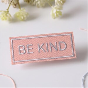 May include: A rectangular fabric patch in a soft peach color featuring the text BE KIND embroidered in light gray thread. The patch has a matching light gray stitched border that frames the lettering, creating a clean and defined rectangular shape.