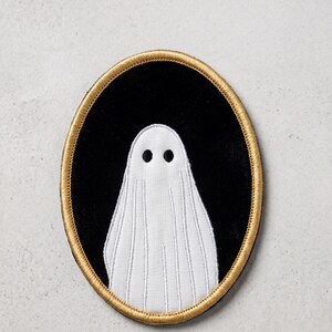 May include: An oval-shaped embroidered patch featuring a white ghost figure with two black circular eyes centered on a solid black background. The entire design is framed by a thick, metallic gold stitched border that defines the oval shape.