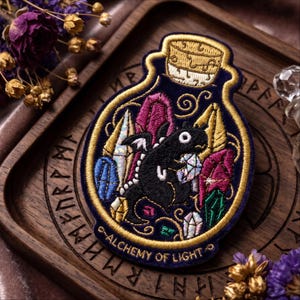 May include: An embroidered patch shaped like a glass bottle containing a black dragon surrounded by colorful crystals. The patch features gold thread borders, a cork stopper, and the text "ALCHEMY OF LIGHT" stitched in gold at the bottom edge.