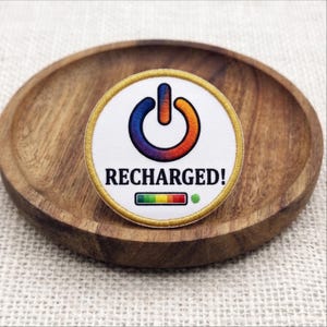 May include: A circular embroidered patch with a white background and a gold border. It features a rainbow-colored power symbol, the text RECHARGED! in black, and a small horizontal battery level indicator bar with a green dot at the end.