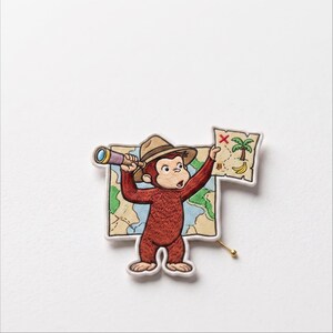 May include: An embroidered patch featuring a brown monkey wearing a tan hat, holding a telescope and a treasure map. The map displays a palm tree, a red X, and yellow bananas. The patch has a white border and a gold-toned metal pin backing.