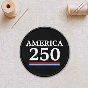 May include: A black circular patch with the text "AMERICA 250" in white. Below the numbers is a horizontal stripe in red, white, and blue. The patch is embroidered with a black border. Sewing supplies are in the background.