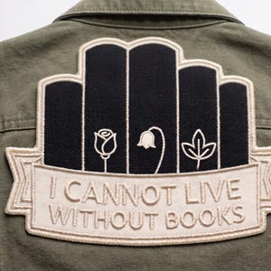 May include: An olive green jacket featuring a large embroidered patch on the back. The patch depicts five black vertical book spines with white line art of a rose, a bellflower, and a leaf, above a banner reading I CANNOT LIVE WITHOUT BOOKS.
