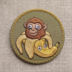 May include: A circular embroidered patch featuring a brown monkey head emerging from a peeled yellow banana. The banana has two small white eyes embroidered on its side. The design is set against an olive green fabric background with a gold border.