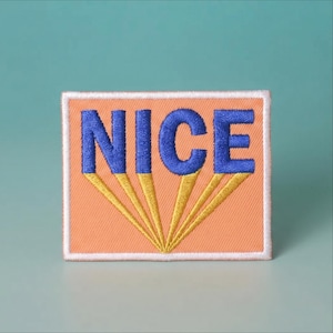 May include: A rectangular embroidered patch featuring the word NICE in bold blue letters. Below each letter, yellow embroidered lines extend downward to a single point at the bottom center. The background of the patch is peach-colored.