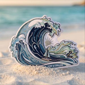 May include: An embroidered patch featuring a large crashing ocean wave in shades of navy, light blue, and white. A small sailboat is positioned at the base of the wave with a sun icon above it. The text SURF & EXPLORE is stitched at the bottom.