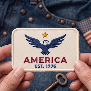 May include: A rectangular embroidered patch with a cream-colored background. The design features a dark blue eagle with wings spread wide, a gold star above the eagle, and the red text AMERICA with the blue text EST. 1776 centered below it.