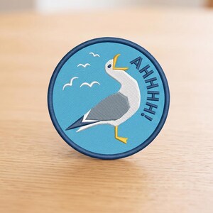 May include: A circular embroidered patch with a light blue background featuring a white and gray seagull with a yellow beak and feet. The bird is depicted with its beak open, and the text AHHHH! is embroidered in dark blue next to it.