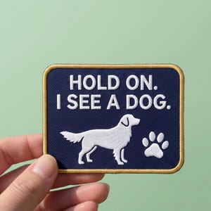 May include: A rectangular embroidered patch with a dark blue background and a gold border. The text reads HOLD ON. I SEE A DOG. in white capital letters. Below the text is a white silhouette of a dog and a white paw print icon.