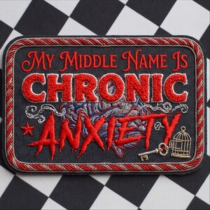 May include: A rectangular embroidered patch with a black background and a red and silver twisted cord border. The text reads MY MIDDLE NAME IS CHRONIC ANXIETY in red and white thread. It features a brain illustration, a small star, a key, and a cage.